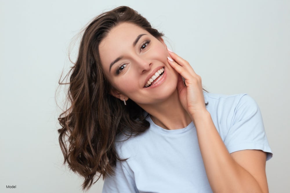 woman in a blue T-shirt smiling and touching her face with her hand.